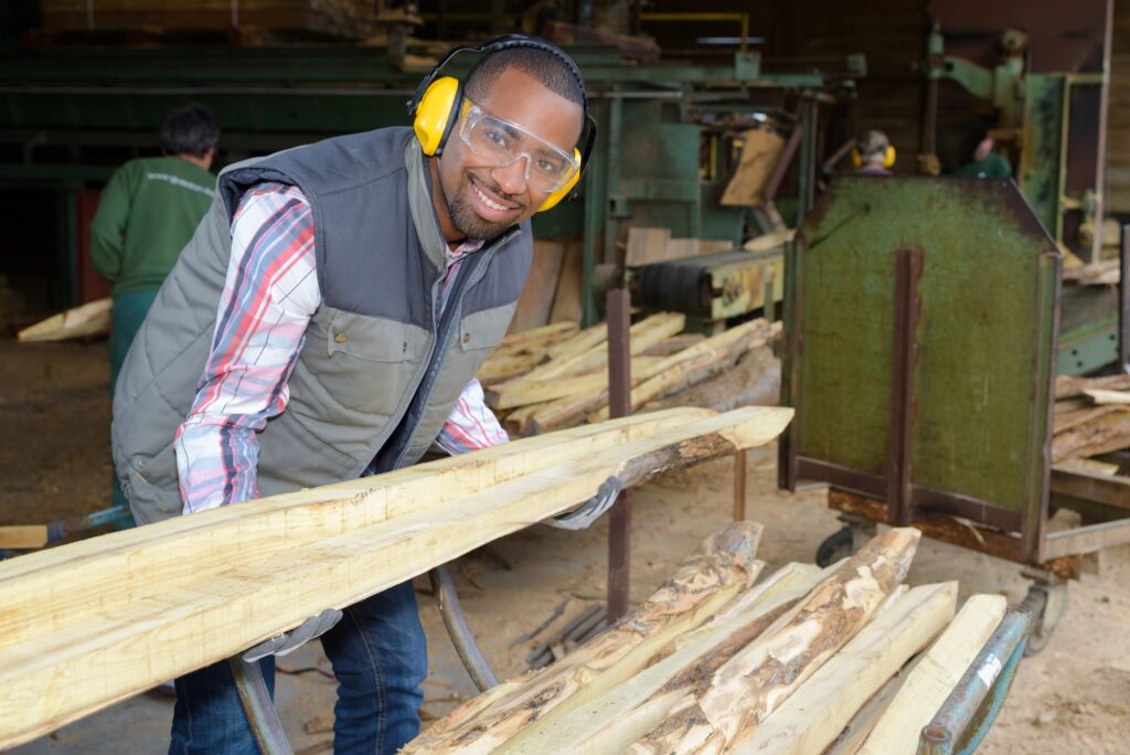 Persoon met veiligheidsbril, gehoorbescherming en handschoenen werkt met houten balken